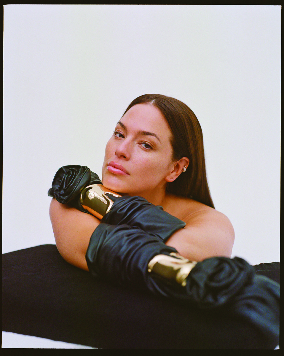 Adult female rests her arms on a surface wearing golden bracelets and black gloves in a studio shot against a white background.