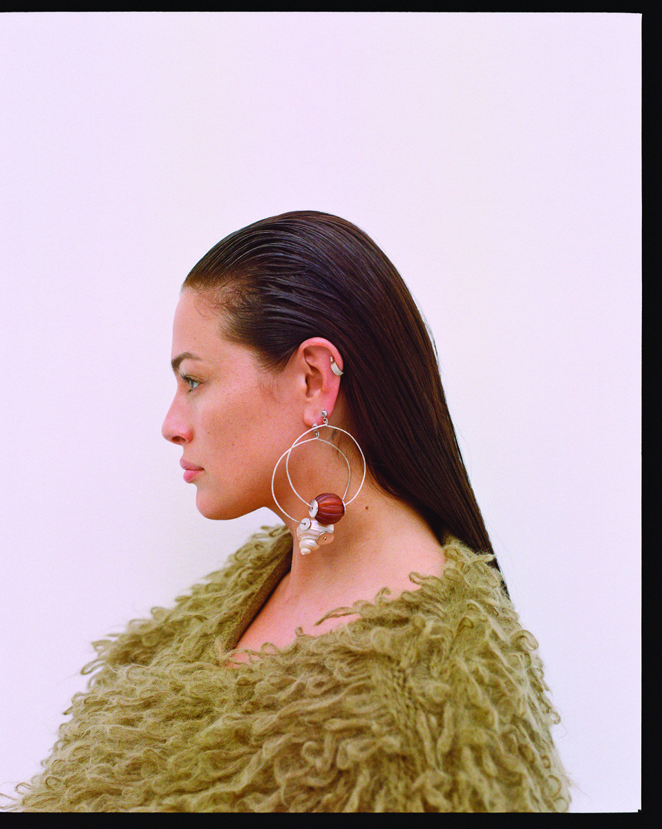 Profile view of a young adult woman with slicked-back hair, large hoop earrings, and a fuzzy olive green sweater against a white background.