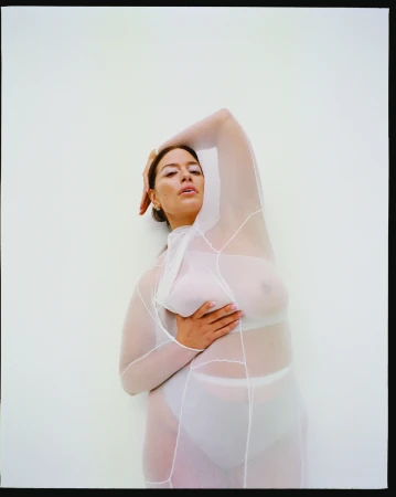 Ashley Graham wearing a sheer white paneled bodysuit posing against a plain white studio background.