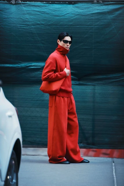 Asian male fashion model in red sweater and wide-leg pants carries a matching bag against a green wall next to a white car.