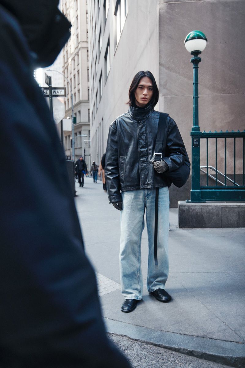 A stylish young Asian man in a black jacket and light-wash jeans stands on a city street with buildings and a subway entrance in the background.