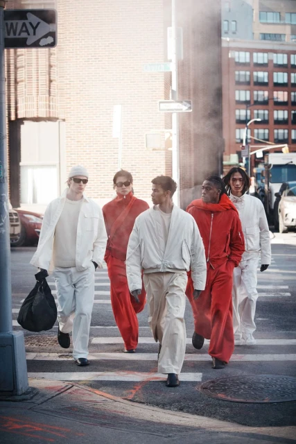 Five people walk across a city street crosswalk. Two wear red jumpsuits, and three wear white or tan jumpsuits. Modern urban scene.