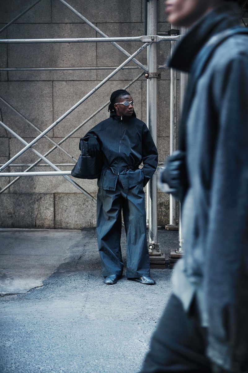 A person stands in an urban environment, wearing a dark blue jumpsuit and sunglasses, with scaffolding and another person visible.