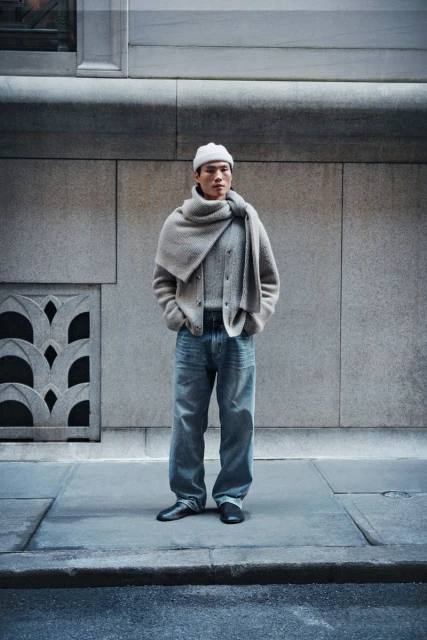 Full-length studio shot of a male model wearing a beige jacket, blue jeans, and a white beanie in front of a building.
