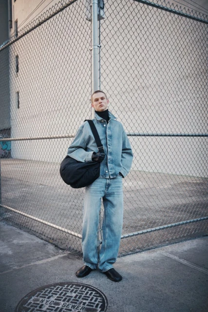 Adult male in a denim jacket and jeans standing in front of a chain-link fence with a black bag hanging over his shoulder in an urban setting.