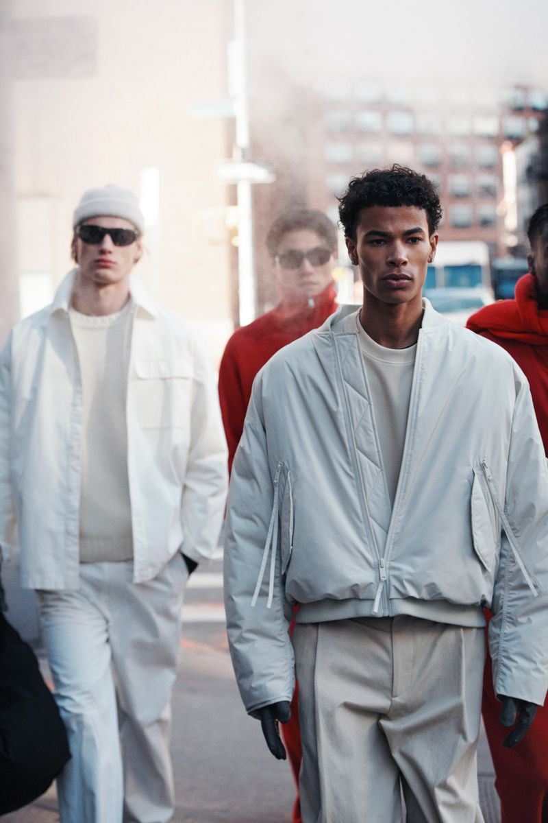 Fashion editorial shot of group of male models walking outdoors wearing bomber jackets and casual street wear in daytime urban environment.