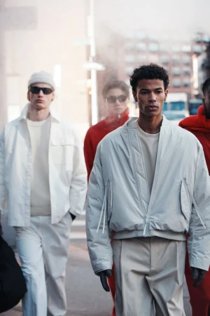 Fashion editorial shot of group of male models walking outdoors wearing bomber jackets and casual street wear in daytime urban environment.