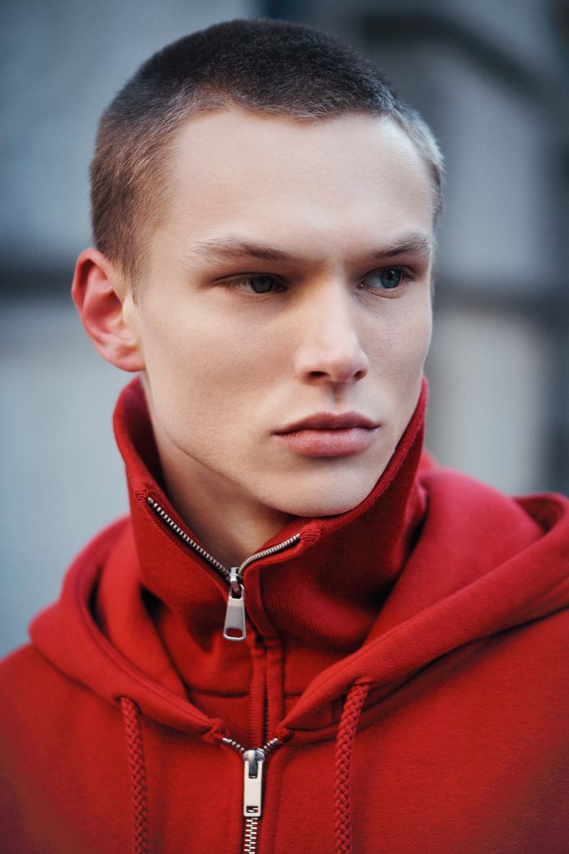 Close-up portrait of a fair-skinned young adult male with a buzzcut wearing a red zip-up hooded sweatshirt, gazing to the side.