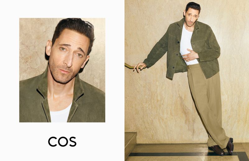 Portrait of actor Adrien Brody wearing an olive jacket and tan pants, leaning against a wall for a fashion editorial for COS.