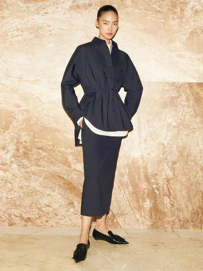 Model in navy blue shirt and matching skirt standing against a polished marble wall for COS SS 2025 campaign.