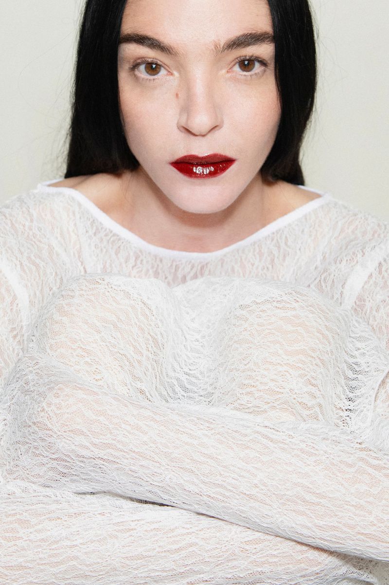 Close-up portrait of model Mariacarla Boscono in a white lace top with dark red lips against a seamless background.