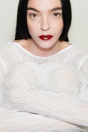 Close-up portrait of model Mariacarla Boscono in a white lace top with dark red lips against a seamless background.