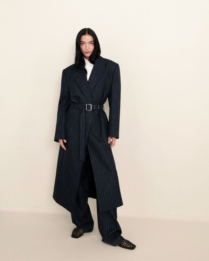 An adult woman in a pinstripe suit coat with a belt and matching trousers posing in a studio setting with a neutral backdrop.