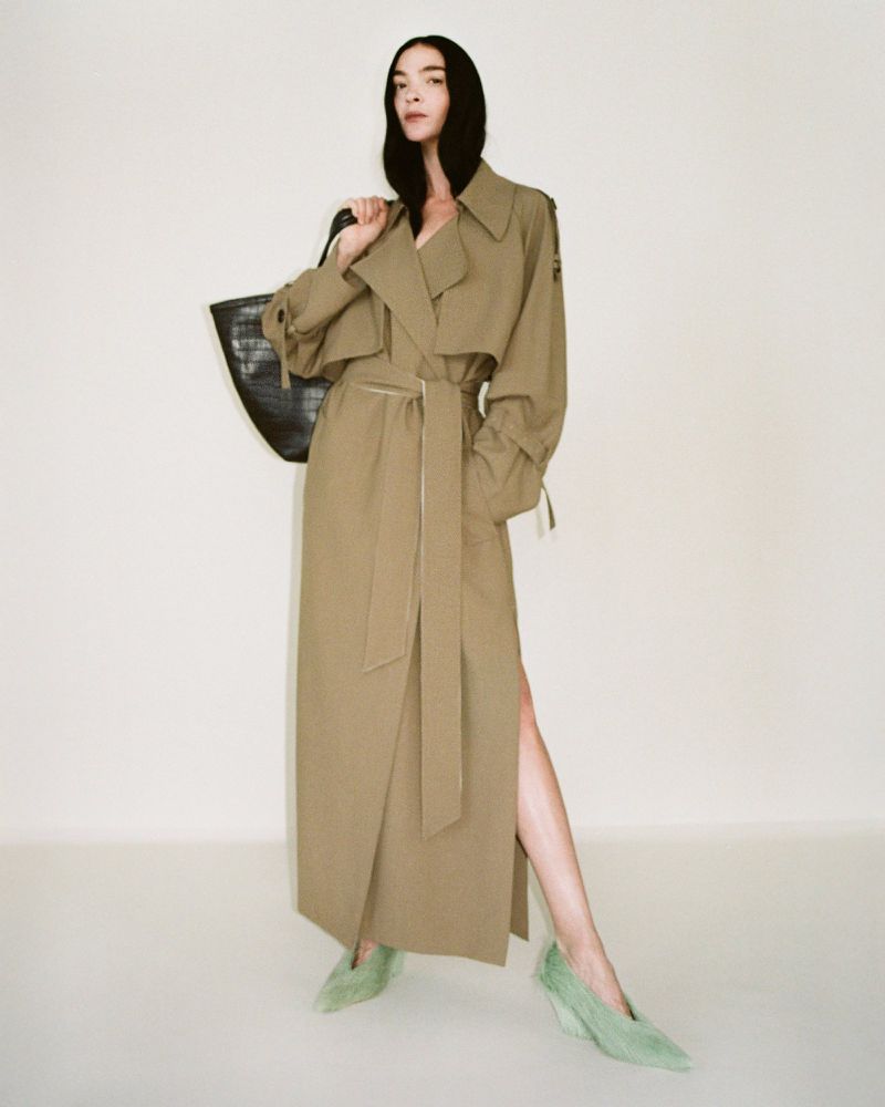 Model wearing a long belted coat and light green heels holding a black handbag against a seamless white background.
