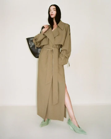 Model wearing a long belted coat and light green heels holding a black handbag against a seamless white background.