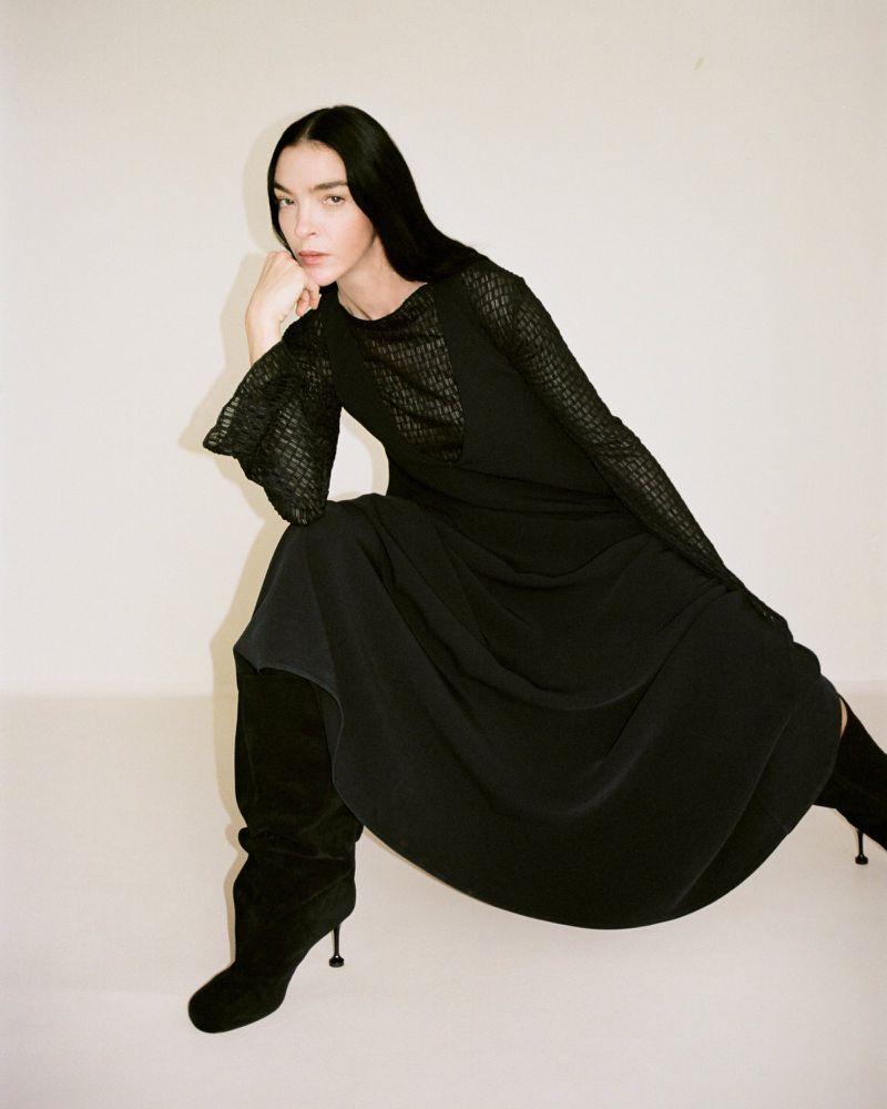 Mariacarla Boscono in a black long-sleeved dress and heeled boots posed against a pale seamless studio background.