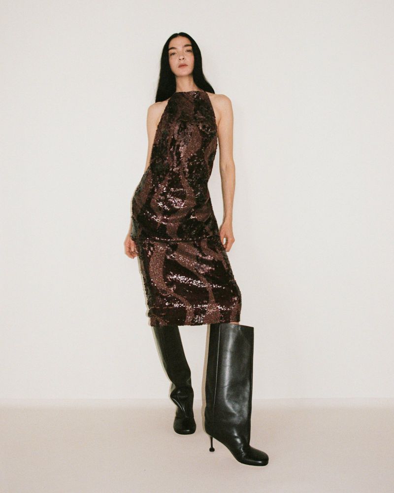 A woman poses in a sparkly dress and knee-high boots against a white background, captured in a full shot fashion studio setting.