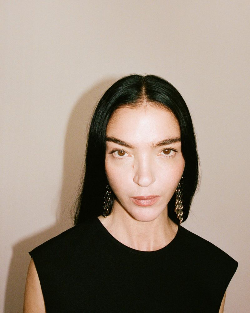 Portrait of model Mariacarla Boscono wearing a black top and statement earrings against a neutral background