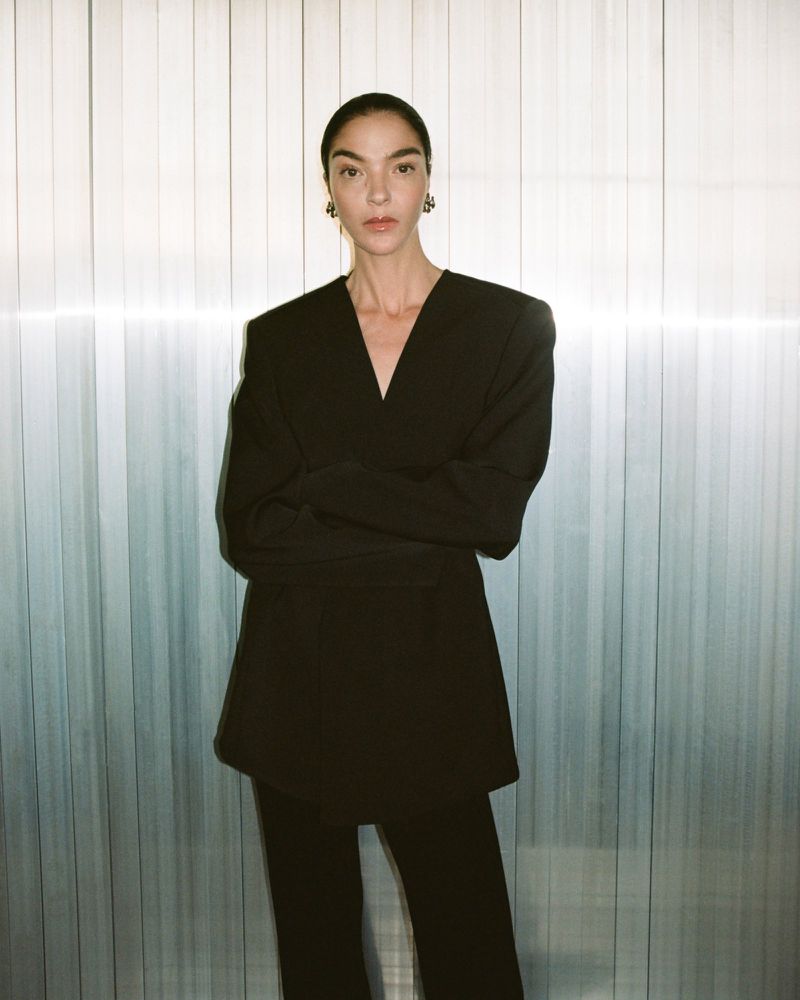 Woman in a dark pant suit, arms crossed, stands against a paneled wall in a fashion portrait with neutral expression.