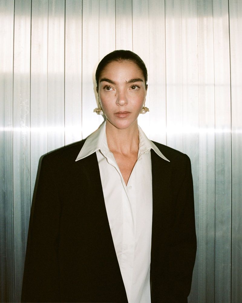Mariacarla Boscono in a black blazer and white shirt against a vertical paneled metallic wall for Camilla and Marc
