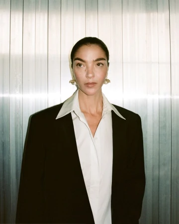 Mariacarla Boscono in a black blazer and white shirt against a vertical paneled metallic wall for Camilla and Marc