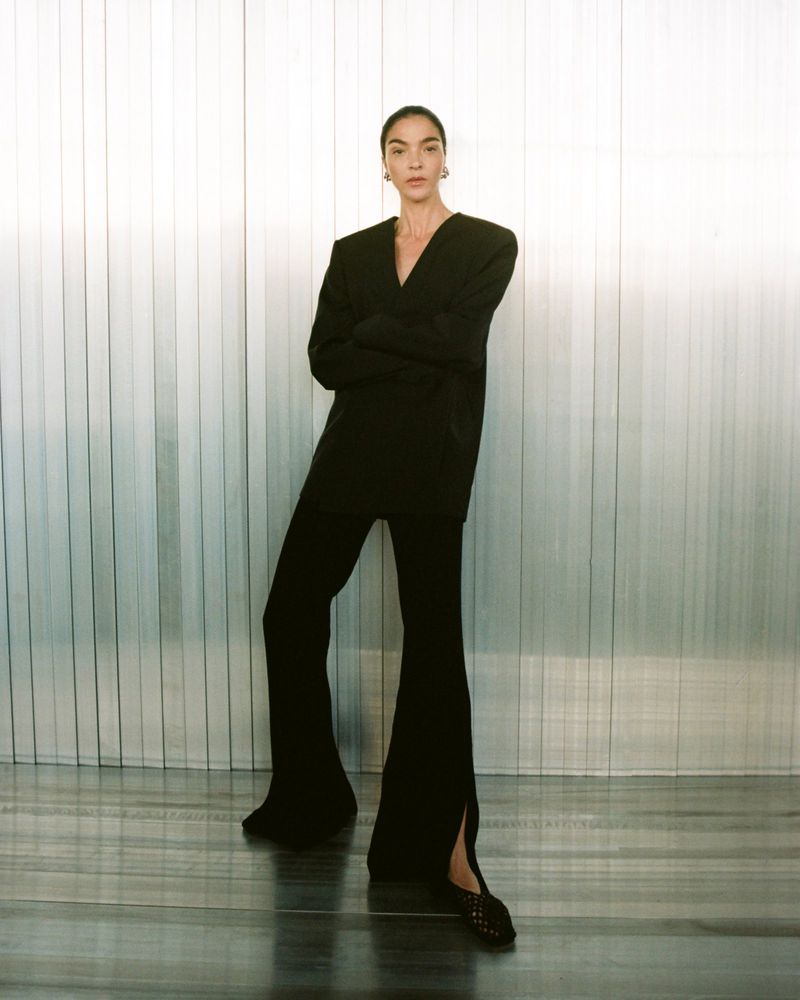 Fashionable adult woman poses in a black blazer and flared pants against a grey-striped wall in a studio setting.