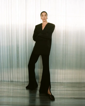Fashionable adult woman poses in a black blazer and flared pants against a grey-striped wall in a studio setting.