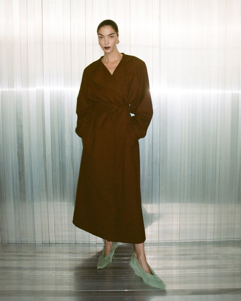 An adult female fashion model poses in a long brown wrap dress, complemented by green shoes, set against a textured silver backdrop.