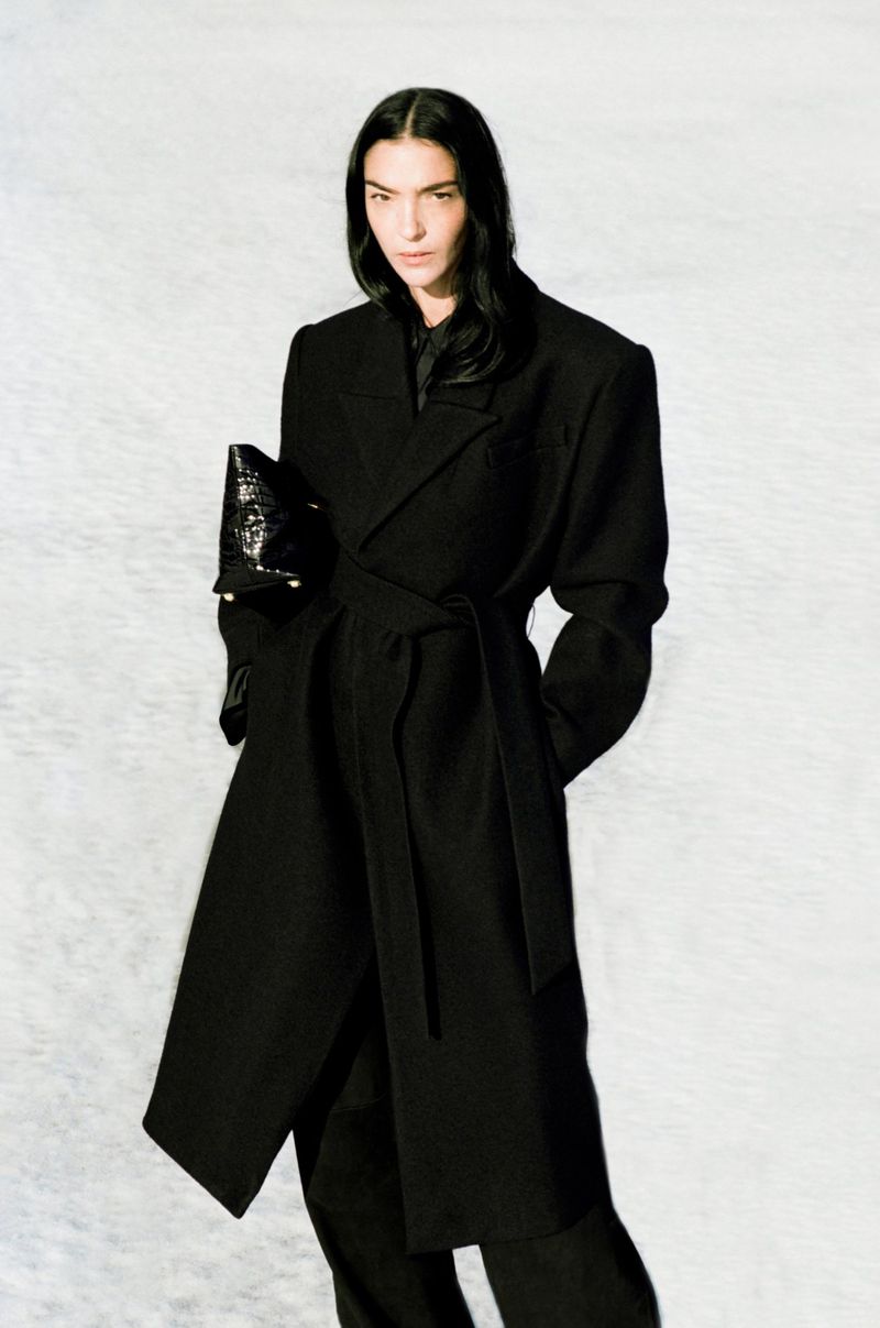 Mariacarla Boscono in a long black belted coat holding a black handbag against a light background for Camilla and Marc S/S 2025 campaign