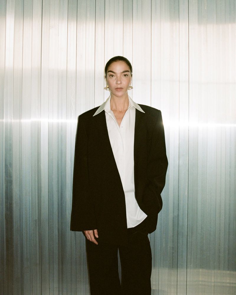 A woman in a black suit and white shirt stands indoors against a textured metal background, posing in a full shot with good lighting.