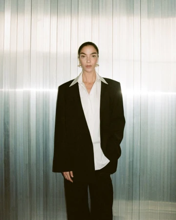 A woman in a black suit and white shirt stands indoors against a textured metal background, posing in a full shot with good lighting.