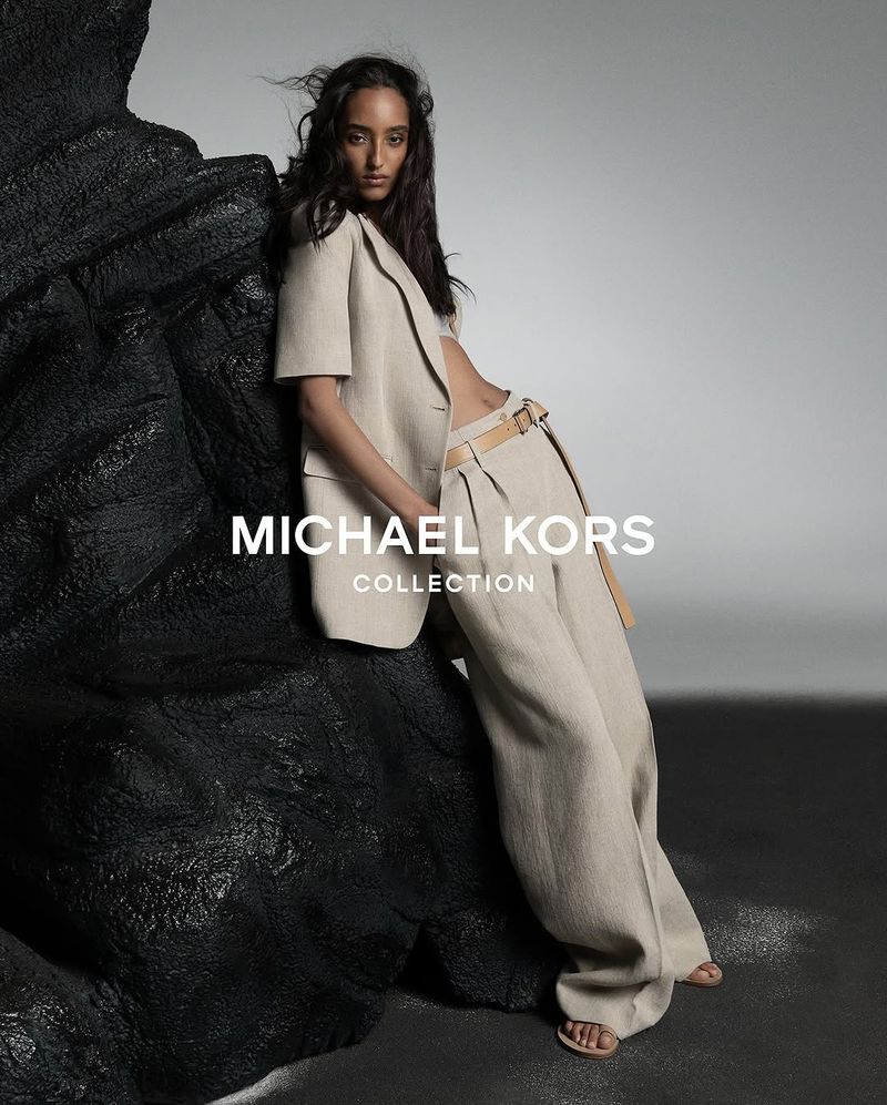 Fashion model in a beige linen suit poses next to a textured black boulder in a studio. Michael Kors Collection ad.