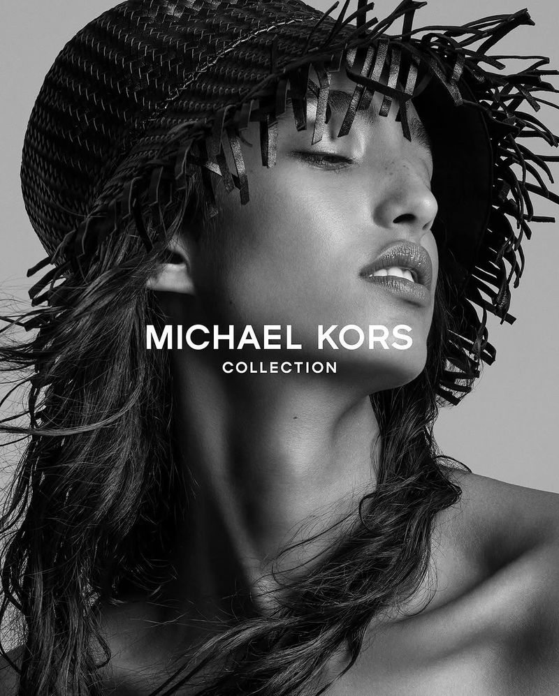 A model with dark hair wears a straw hat with fringe in a black and white fashion portrait for Michael Kors Collection.