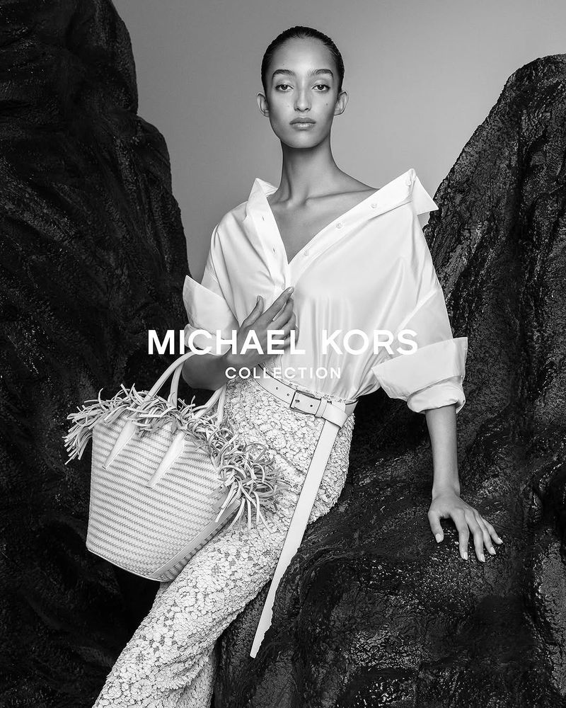 Fashion shot of a model posing in a white shirt and lace pants, accessorized with a woven straw bag, set against a dark rocky backdrop.