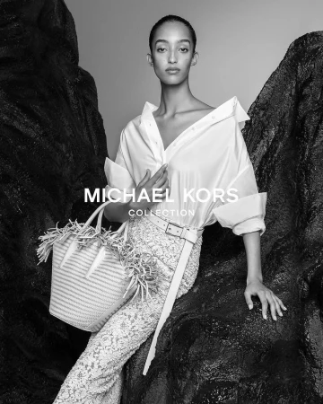 Fashion shot of a model posing in a white shirt and lace pants, accessorized with a woven straw bag, set against a dark rocky backdrop.