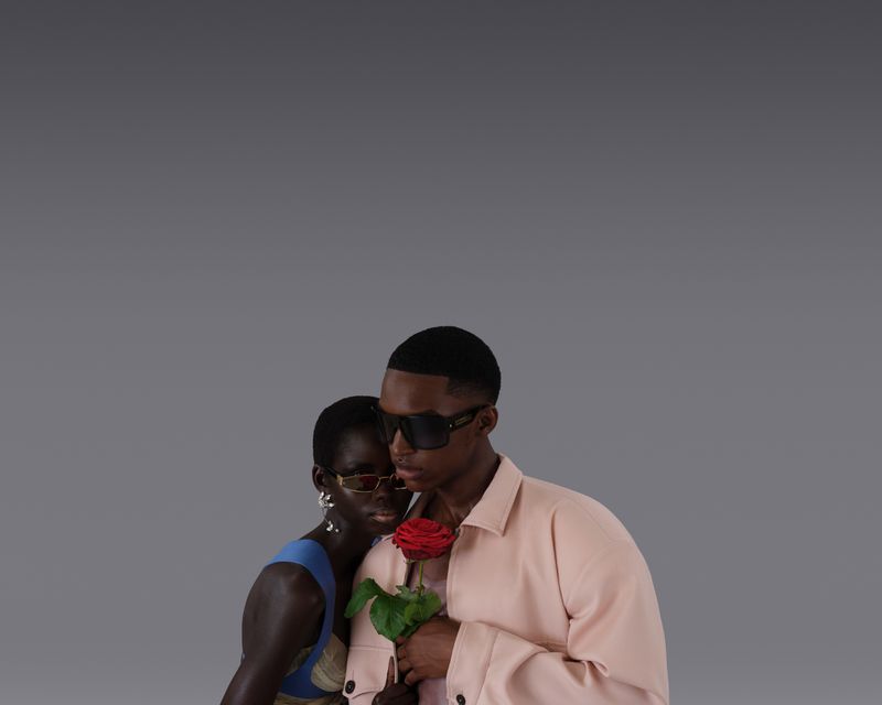 Fashion shot of an African American woman and man in sunglasses, with the man holding a red rose against a solid gray backdrop.