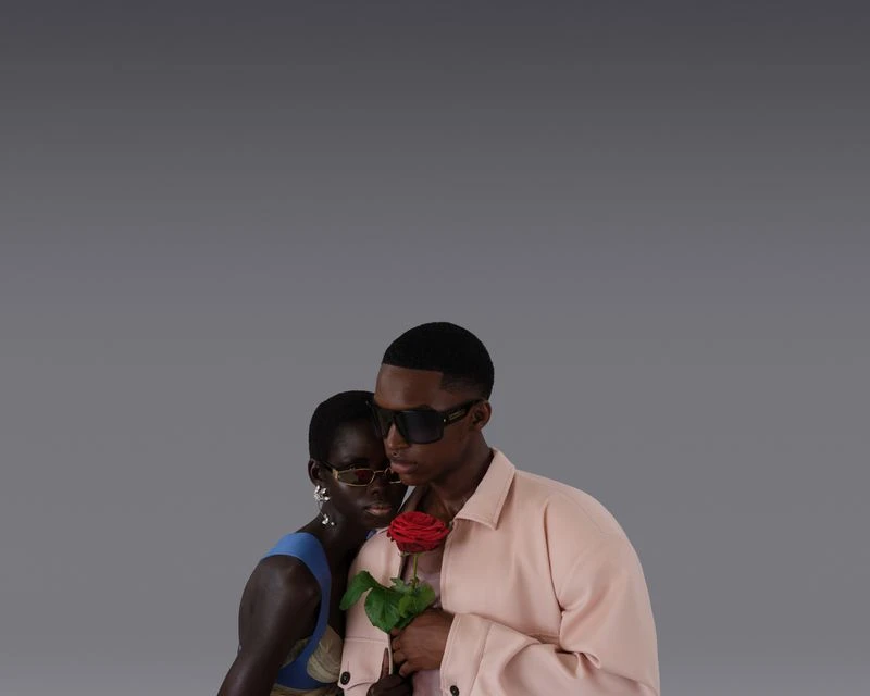 Fashion shot of an African American woman and man in sunglasses, with the man holding a red rose against a solid gray backdrop.