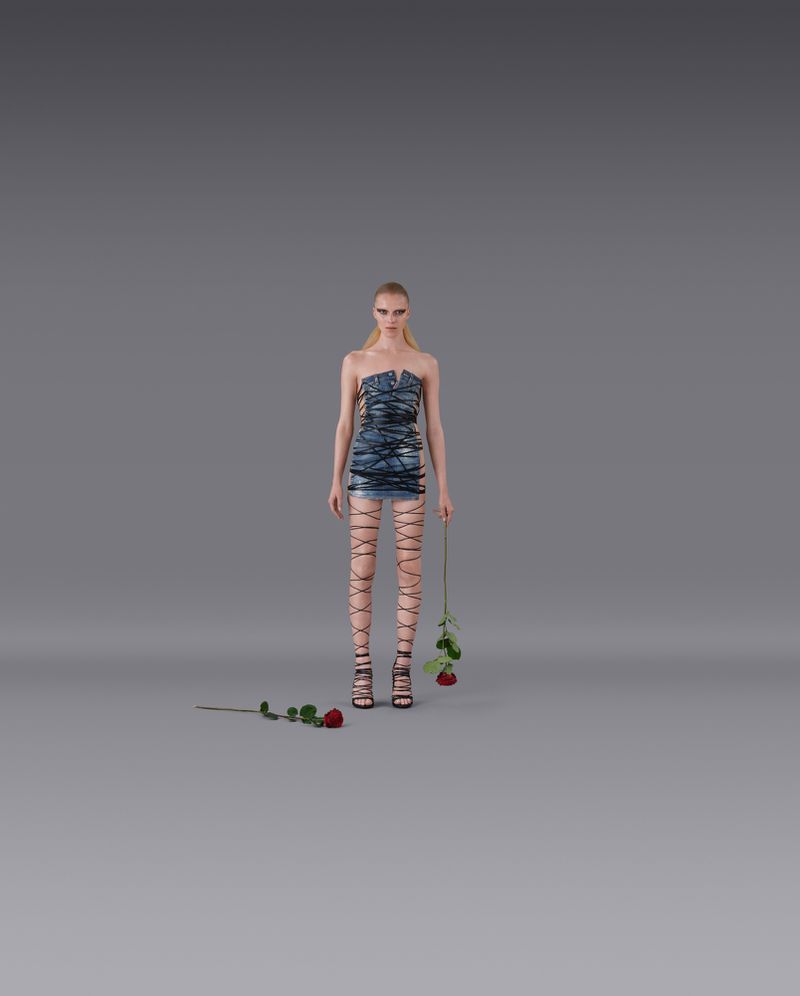 Fashion model in a blue dress and strappy heels poses in a studio setting with roses, against a neutral gray backdrop, medium full shot.