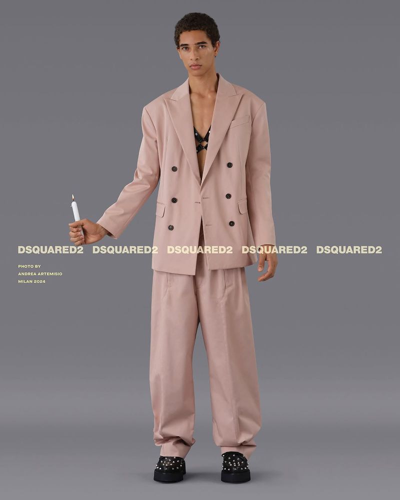 Fashion shot of a young adult model in a pink suit holding a candle, posing in a studio against a grey backdrop with brand logos.