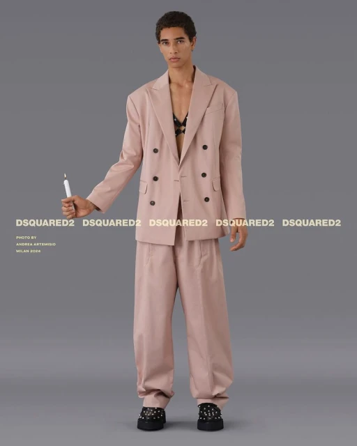 Fashion shot of a young adult model in a pink suit holding a candle, posing in a studio against a grey backdrop with brand logos.
