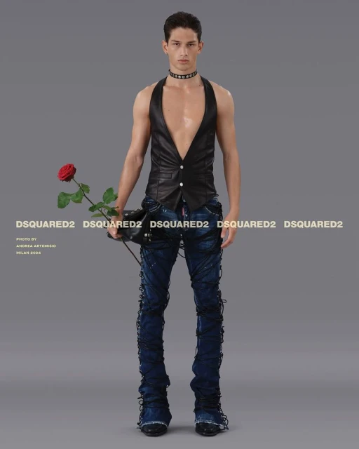A stylish male model poses in a black vest, blue jeans with decorative ties, and a choker, holding a red rose against a grey backdrop.