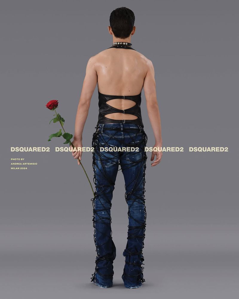 Back view of a slender person in dark halter top and laced jeans holding a rose, studio lit against a gray background for a fashion shoot.
