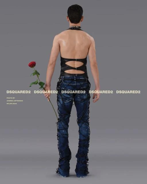 Back view of a slender person in dark halter top and laced jeans holding a rose, studio lit against a gray background for a fashion shoot.