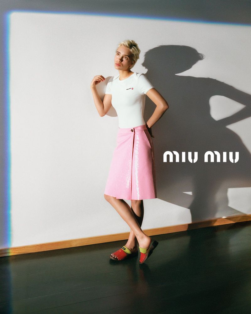 Fashion editorial shot of a woman posing in a white shirt and pink skirt next to the Miu Miu logo in a studio with moody lighting.
