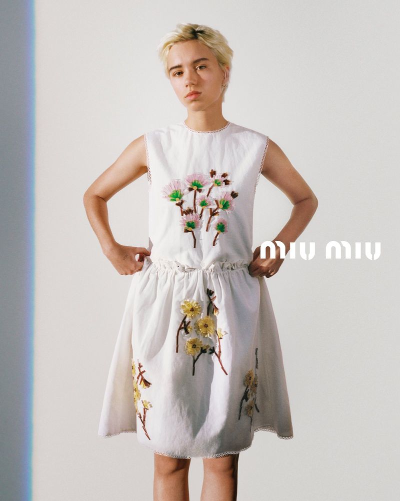 A young, fair-skinned model poses in a white dress embroidered with floral patterns against a bright, minimalist backdrop, showcasing fashion and style.