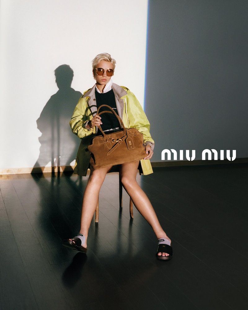 A blond woman is seated in a photo studio, wearing a raincoat, sunglasses, and sandals, holding a tan suede handbag with Miu Miu logo.