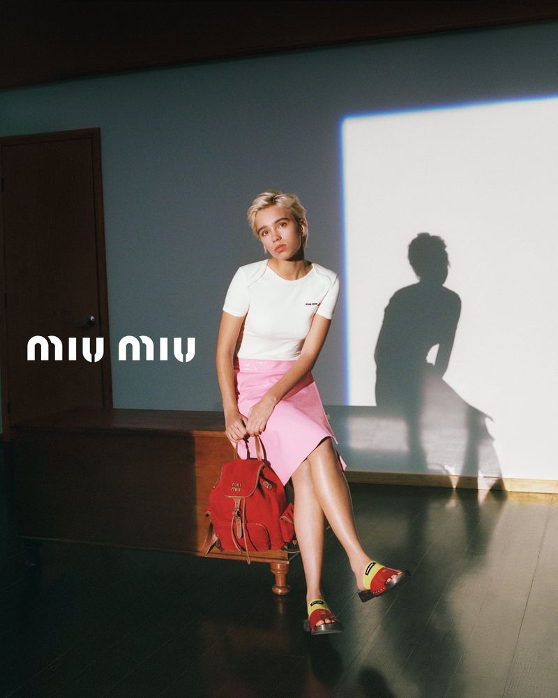 Young woman in a pink skirt sitting in a light-filled room, holding a red backpack, wearing patterned sandals. Miu Miu logo is on the wall.