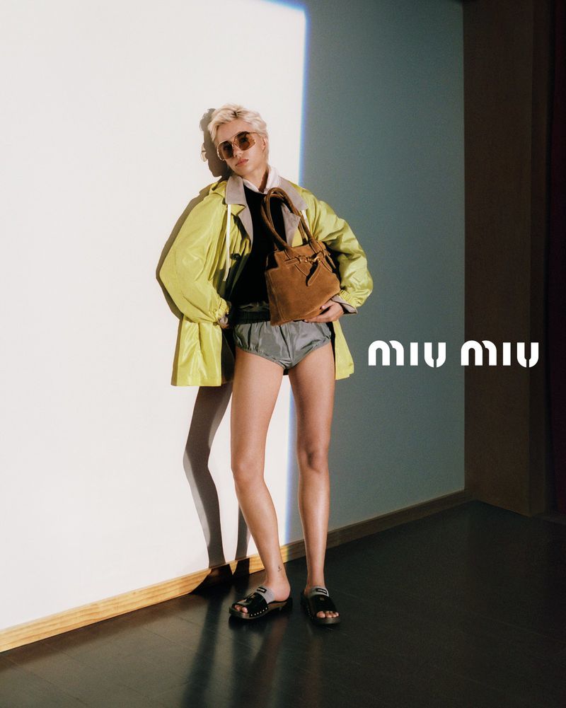 A person wearing a neon jacket, shorts, and sandals poses indoors with a brown bag, standing against a white wall with the Miu Miu logo.