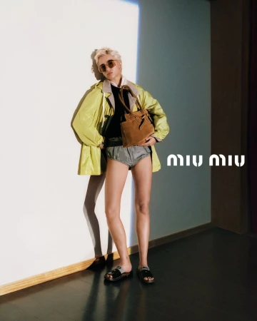 A person wearing a neon jacket, shorts, and sandals poses indoors with a brown bag, standing against a white wall with the Miu Miu logo.