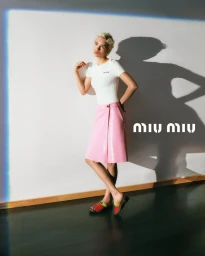 Model with blonde hair wearing a white t-shirt and pink skirt against a plain wall with a sharp shadow cast behind her.